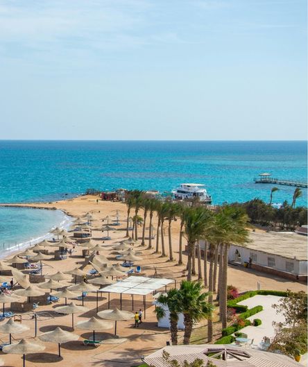 Sandstrand eines Urlaubsresorts am Roten Meer mit Palmen, Reet-Sonnenschirmen und Liegen, im Hintergrund türkisfarbenes Wasser und ein Boot. Sandstrand eines Urlaubsresorts am Roten Meer mit Palmen, Reet-Sonnenschirmen und Liegen, im Hintergrund türkisfarbenes Wasser und ein Boot.
