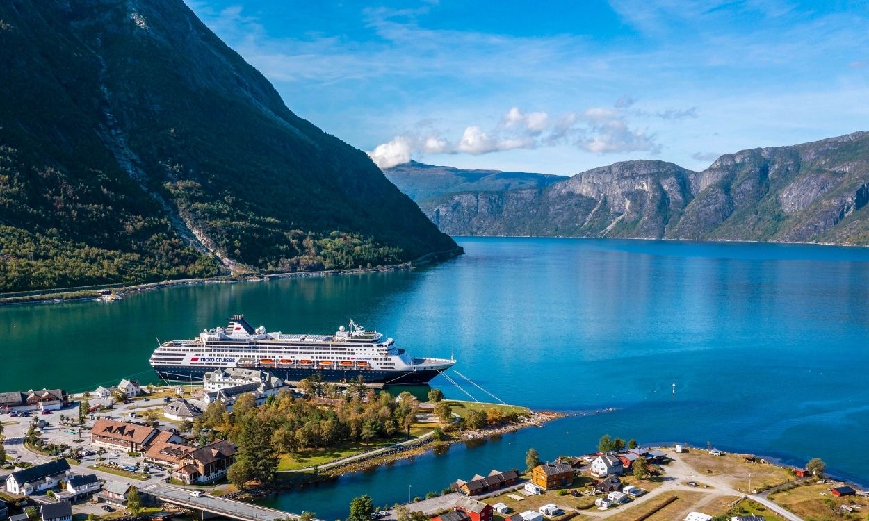 Kreuzfahrtschiff im tiefblauen Fjord von Geiranger, Norwegen, umgeben von hohen, bewaldeten Bergen und einem kleinen Dorf im Vordergrund. Kreuzfahrtschiff im tiefblauen Fjord von Geiranger, Norwegen, umgeben von hohen, bewaldeten Bergen und einem kleinen Dorf im Vordergrund.